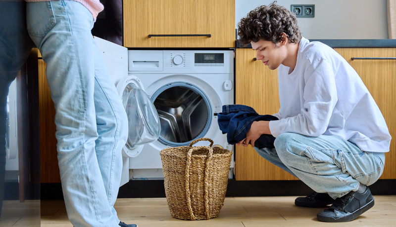 Hands loading mixed, colorful clothes into the front-loading washing machine drum, suggesting a large or varied laundry load best handled by professionals.