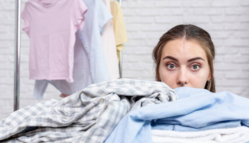 A woman with wide eyes peeking over a huge pile of clothes, symbolizing the feeling of being overwhelmed and stressed by managing complicated laundry at home