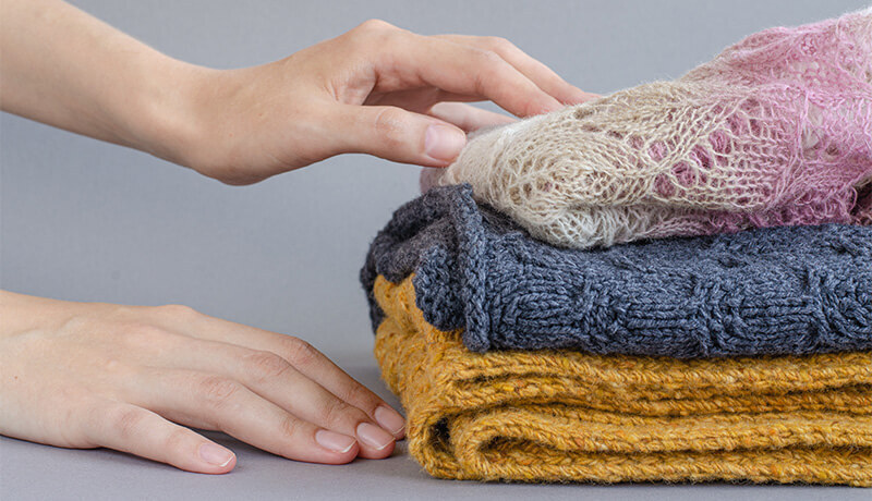 A person’s hands gently touching a stack of folded, high-quality cashmere sweaters in cream, charcoal grey, and mustard yellow.