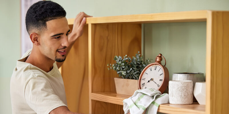 A young man smiling while dusting a wooden bookshelf, demonstrating a routine task from a household chore list to keep the home organized.
