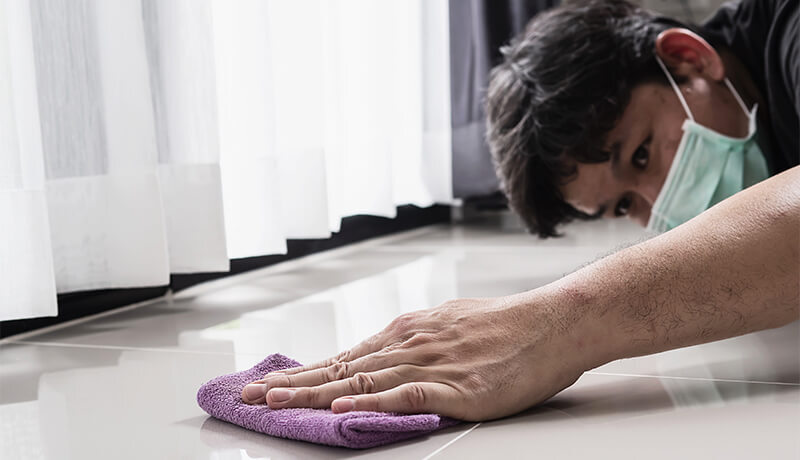 A person wearing a protective face mask meticulously hand-scrubbing a tile floor, illustrating the deep attention to detail found in professional home cleaning services.