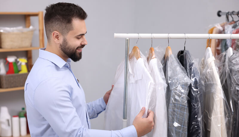 A smiling professional holding two garments in clear protective plastic covers, illustrating the final step of the dry cleaning process for new customers.