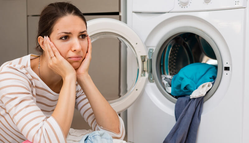 A frustrated woman sitting in front of an open front-load washing machine with clothes spilling out, illustrating the common mistake of overloading the drum.