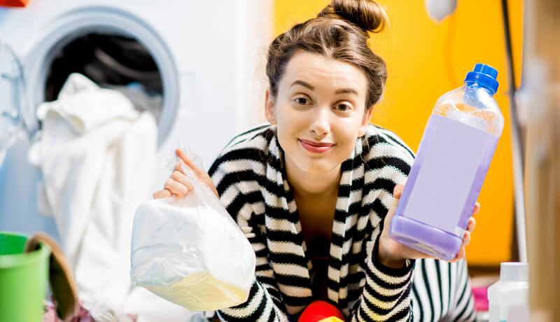 A woman in a striped shirt holding a bottle of purple liquid detergent and a bag of white powder, illustrating the choice between different specialized cleaning agents used in professional garment care.