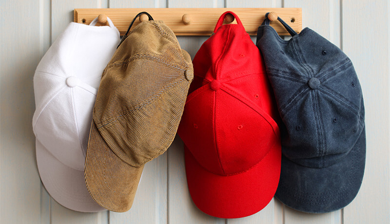 A close-up of four different ball caps—white, corduroy tan, bright red, and faded navy—hanging on a wooden rack, illustrating the need to check fabric types before washing.
