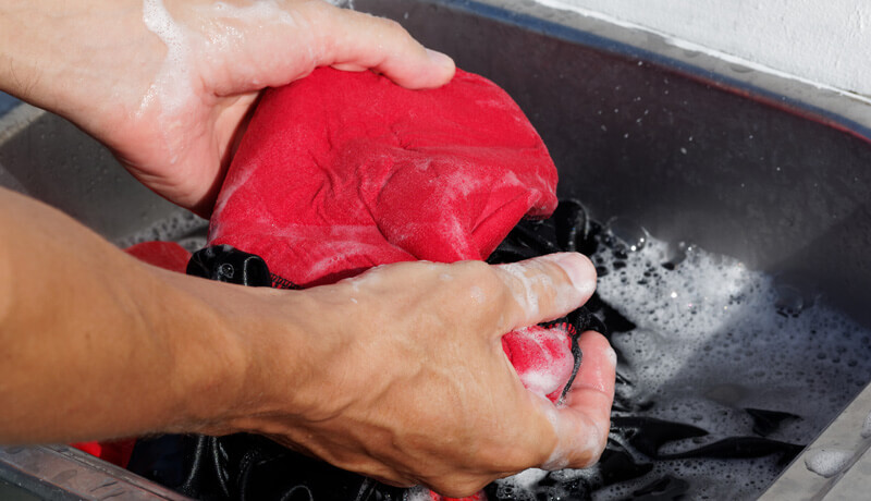 Close-up of a professional technician's hands gently hand-washing a bright red garment in a soapy basin, illustrating the careful manual process used to clean delicate items like ball caps without damaging their structure.