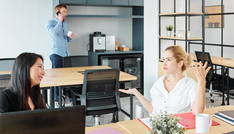Two professional women engaging in a friendly and focused conversation at a clean, modern office desk, illustrating how a clutter-free environment fosters positive employee morale and collaboration.