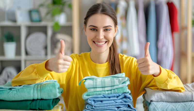 A smiling woman in a yellow sweater giving a double thumbs-up behind perfectly folded stacks of colorful towels, illustrating total satisfaction with professional laundry results.