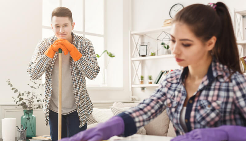A young man in orange gloves leaning on a mop while watching a colleague clean a bright, modern living room, illustrating the teamwork and efficiency behind professional home maintenance services.