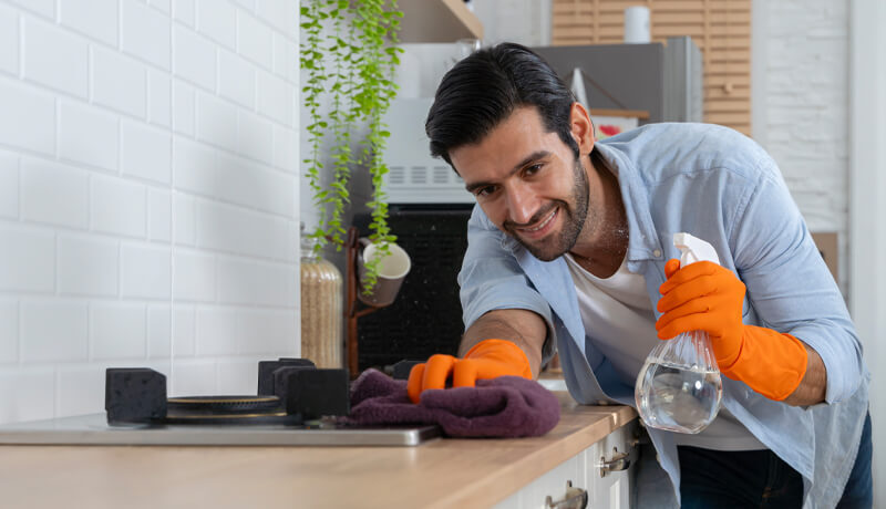 A man wearing orange protective gloves scrubbing a wooden kitchen countertop with a purple cloth and spray bottle, illustrating the physical labor involved in detailed home maintenance.