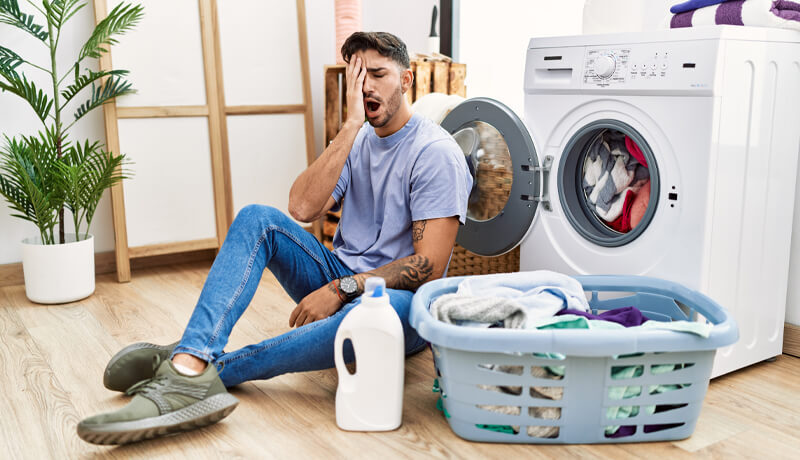 A man sitting on the floor next to a washing machine with his hand over his face in a gesture of frustration, representing the regret of forgetting to empty pockets before starting a load.