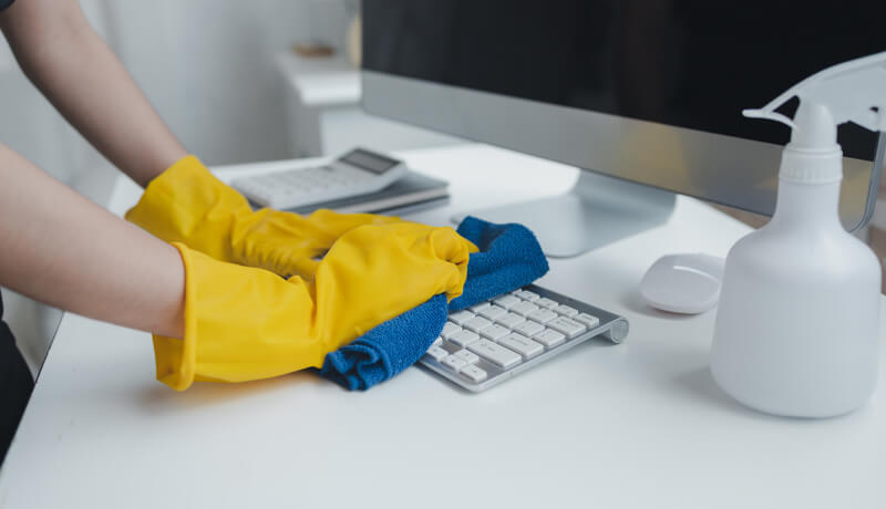 A close-up of a person wearing yellow protective gloves using a blue microfiber cloth to carefully clean a white computer keyboard, illustrating the specialized care needed for sensitive office electronics.