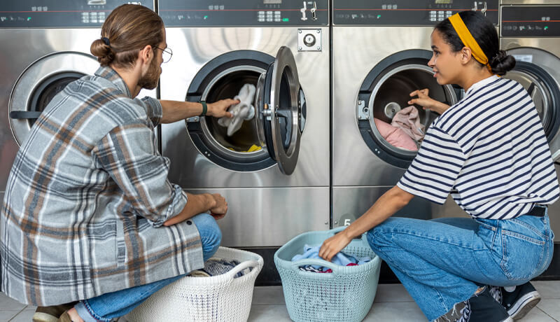 Two people loading clothes into large, industrial-grade stainless steel washing machines at a professional laundromat, representing the high-capacity cleaning required for heavy loads.