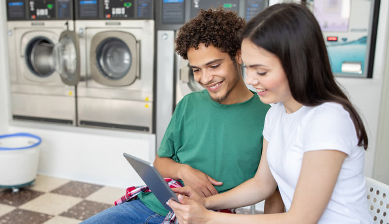 A young couple smiling while using a tablet to schedule a laundry pickup in a modern laundromat, representing the ease of ByNext's digital booking system.