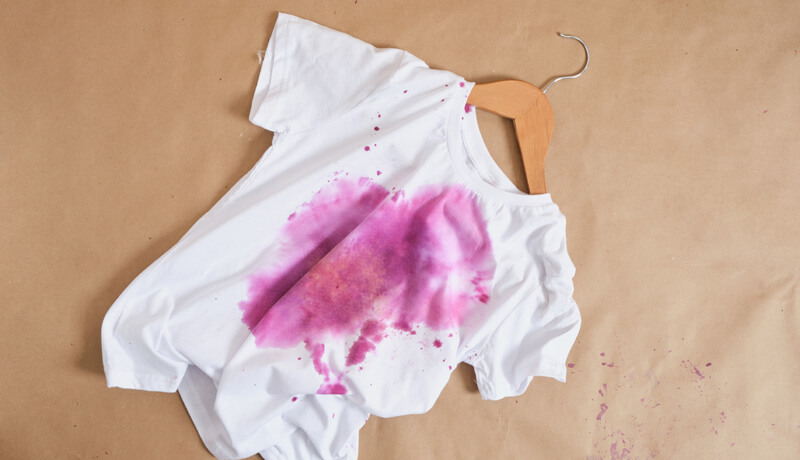 A white t-shirt on a wooden hanger showing a large purple dye stain across the chest, illustrating the damaging effects of color bleeding when clothes are not washed correctly.