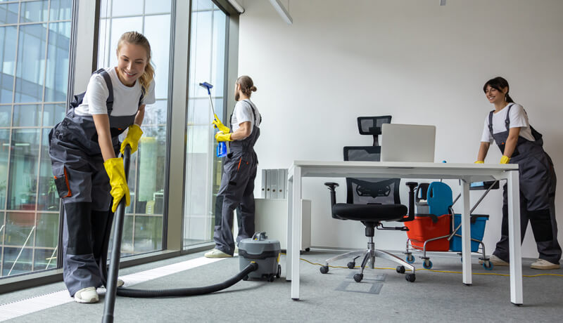 A professional cleaning crew in grey overalls and yellow gloves vacuuming and cleaning windows in a modern, bright office space.