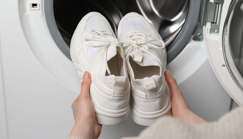 Hands holding a pair of white sneakers in front of an open washing machine door, illustrating a common but risky home cleaning method.