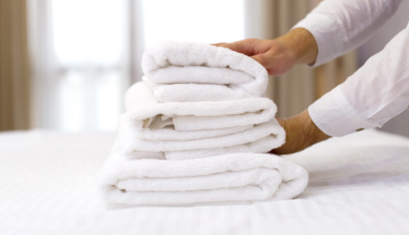 Close-up of a staff member in a crisp white shirt placing a perfectly folded stack of fresh white towels onto a hotel bed, demonstrating ByNext's commitment to high-standard guest room presentation.